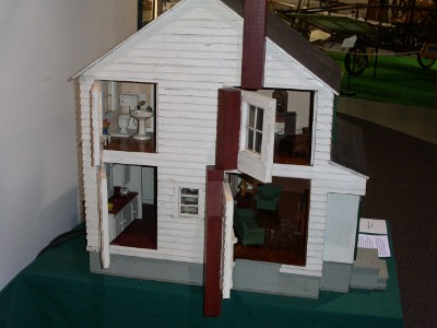 [Two story four room dollhouse with white siding. Part of the siding in four places opens as a door to show four rooms. A bathroom is in the upper left. A kitchen in the lower left. The door, with a window in it, is partially closed on the upper right. The lower right appears to be a livingroom.]